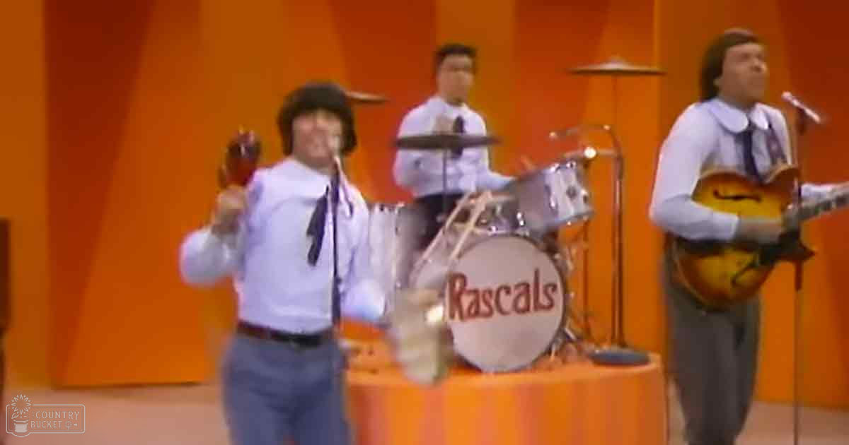 A musical performance scene with performers on stage. The setting includes a drum set labeled 'Rascals', microphones for each performer, and an orange backdrop suggesting it's part of a television show or music video from the 1960s-70s era.