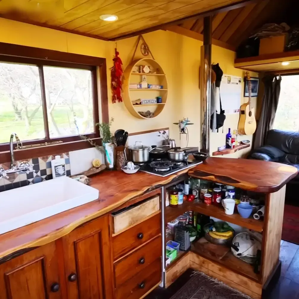A cozy kitchen setup in a small living space with wooden countertops, modern faucet, gas stove, shelves filled with kitchen items, decorative objects on a wall-mounted shelf, and a guitar hanging beside the window showing trees outside suggesting spring or summer.