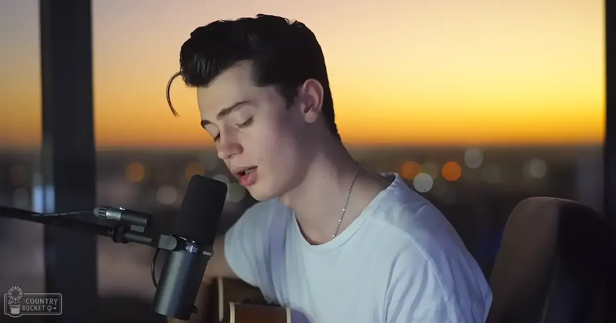 A young adult male sings into a microphone with a cityscape at sunset behind him, wearing a light blue t-shirt in an indoor recording space.