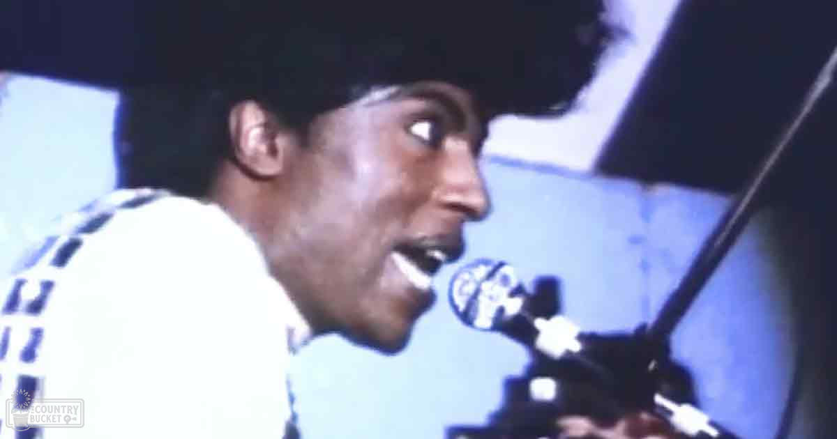 A young adult male wearing a white t-shirt with checkered patterns singing into a microphone in what appears to be a recording studio setting.