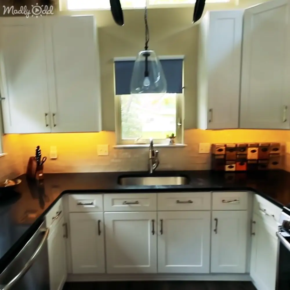 Residential kitchen interior with white cabinets, black countertop, stainless steel sink, blue blinds allowing natural light in, pendant light fixture and a small decorative item near the window.