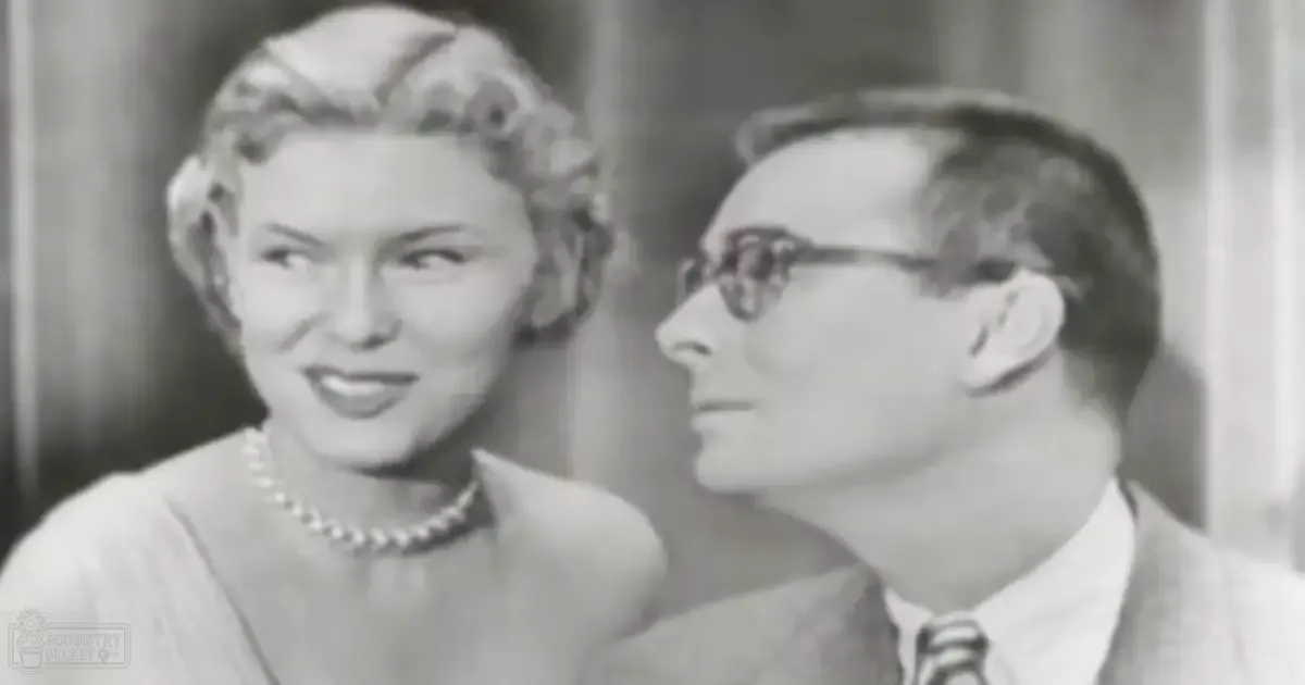 Adult female wearing a light-colored dress or blouse with a pearl necklace indoors next to an adult male in a suit and glasses, natural light from the side, curtain backdrop.