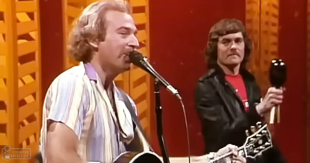 An adult male singer wearing a striped short-sleeved shirt performs into a microphone with another individual playing an instrument, suggesting a live music performance.