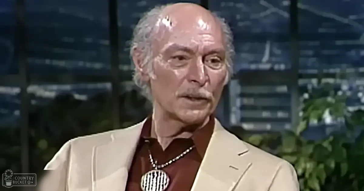 An older adult male wearing a tailored, light-colored blazer over a collared shirt with a pendant necklace stands against a studio backdrop with cityscape windows and greenery at night.