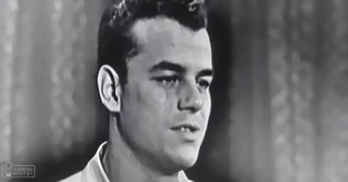 Young adult male wearing off-white collared shirt, mid-20th century style, in an indoor setting with curtains suggesting a home or stage backdrop.