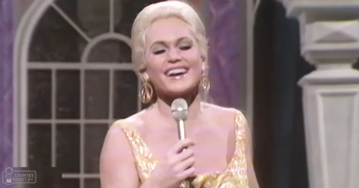 Adult female performer wearing a sleeveless gold dress with intricate patterns and sequins, standing indoors near a window with a grid pattern.