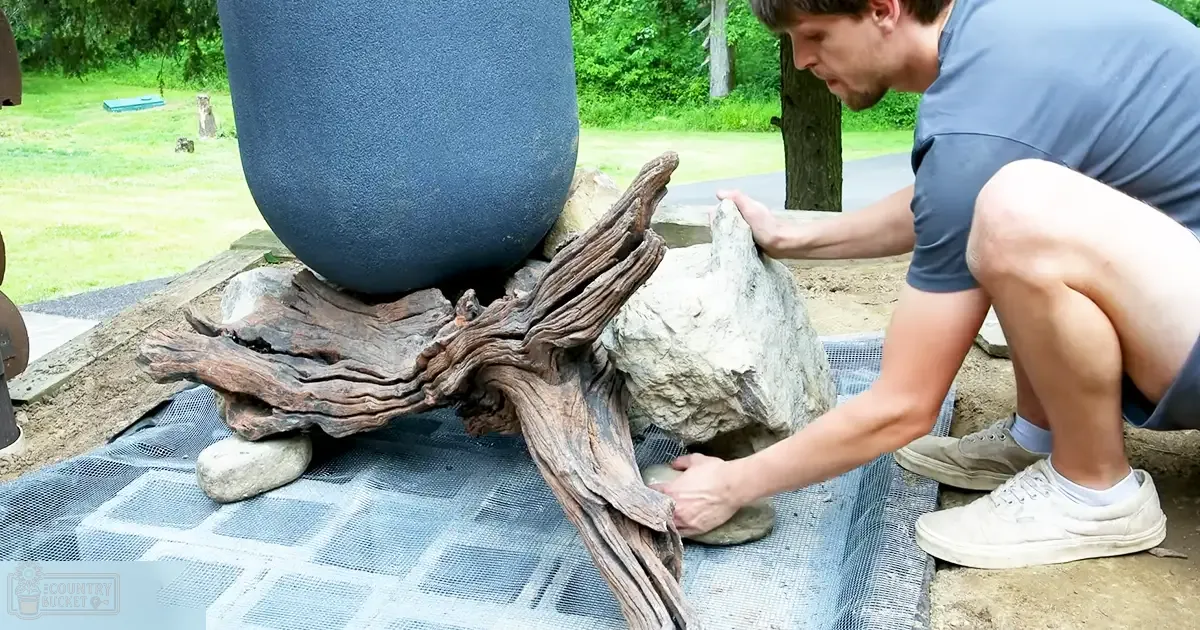 An adult male wearing a blue, relaxed-fit crew-neck t-shirt and shorts, arranging driftwood and rocks in an outdoor garden or park setting with lush greenery.