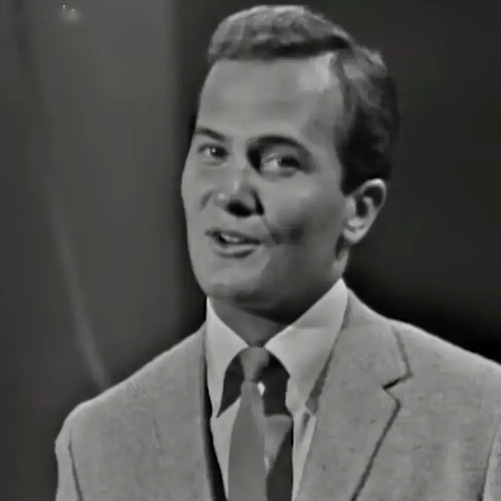 A young adult male in a tailored light gray suit, white collared shirt with dark tie against an indoor backdrop.