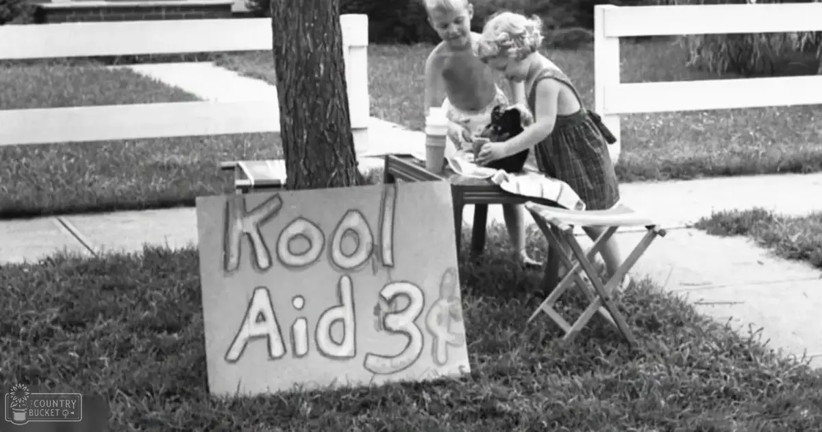 A young girl selling Kool Aid in front of her suburban home, accompanied by a small dog with brownish-gray coat and dark patches.
