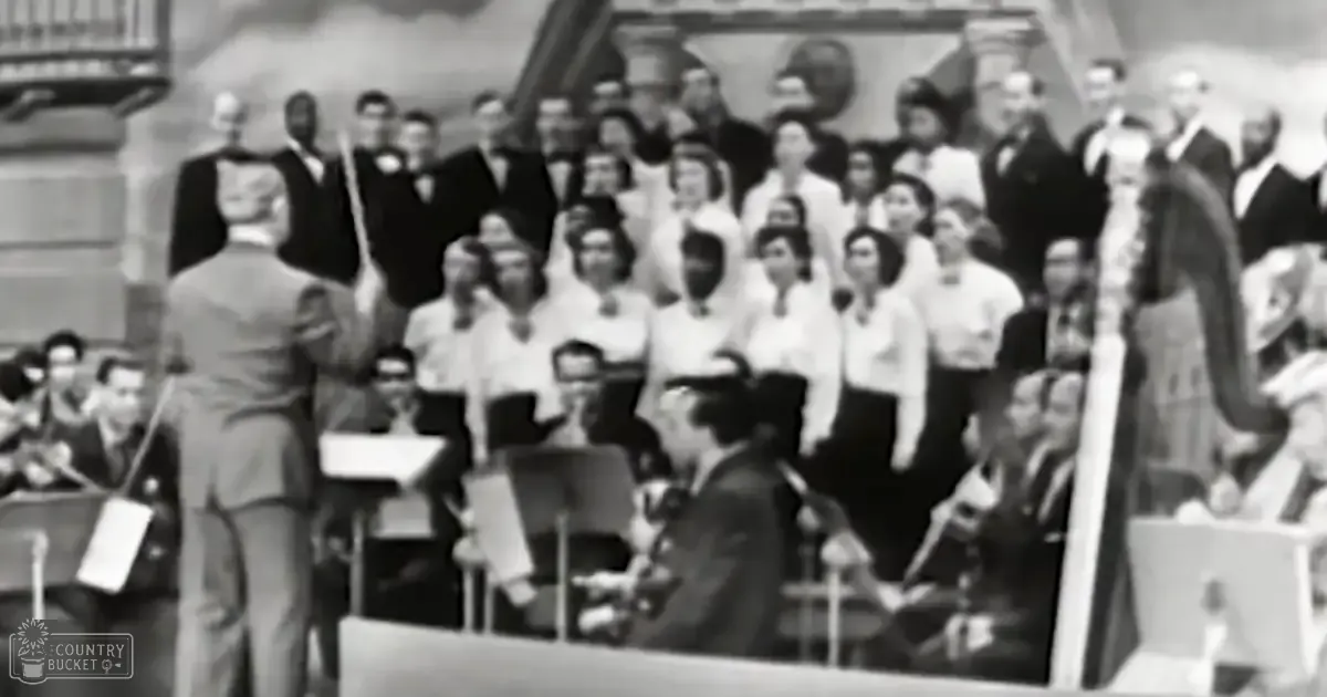 A conductor leads an orchestra and choir in a formal musical performance within the grand architecture of a concert hall, with sheet music stands and stringed instruments visible.