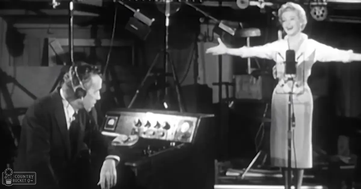 A middle-aged to older adult female in a white dress with long sleeves stands next to an adult male singer wearing a dark suit jacket and white shirt, both positioned near recording studio equipment like microphones and a mixing console.