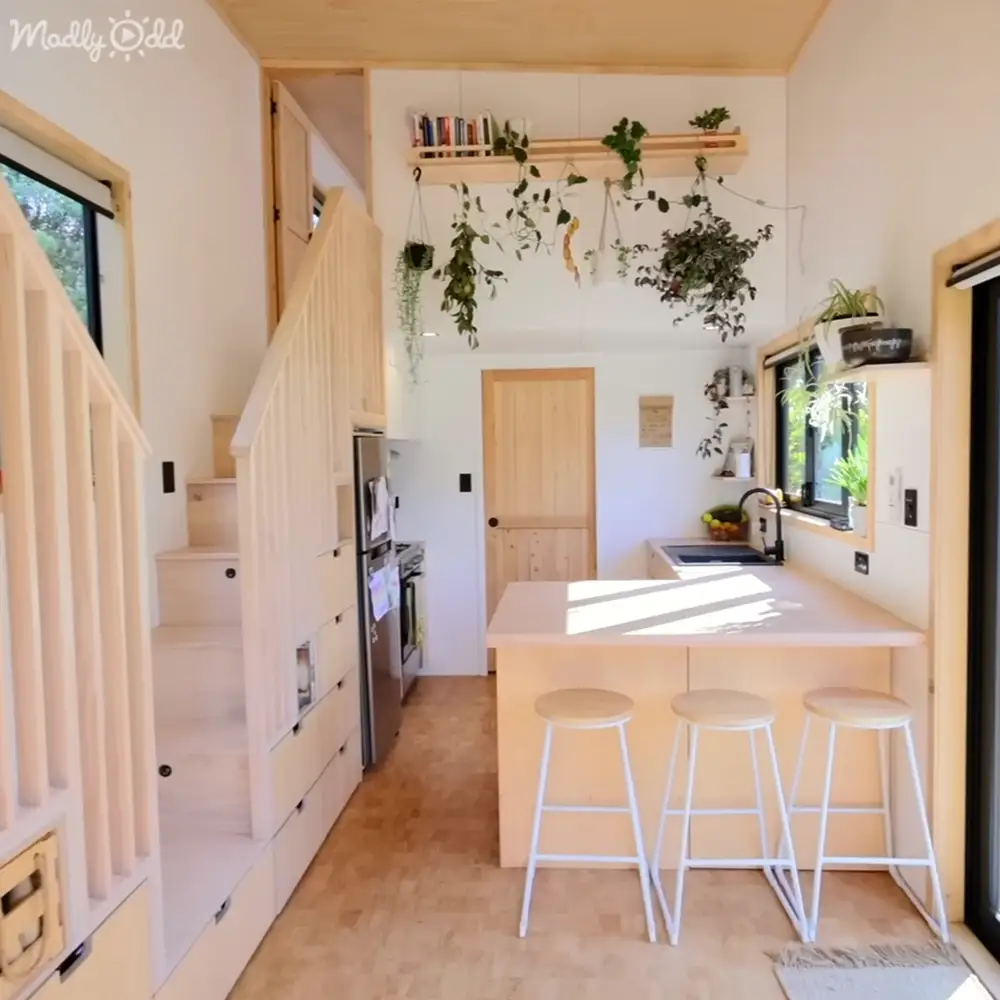A modern kitchen with a wooden staircase leading up to it, with hanging plants on the wall above a white countertop with three stools underneath, set indoors during daytime.