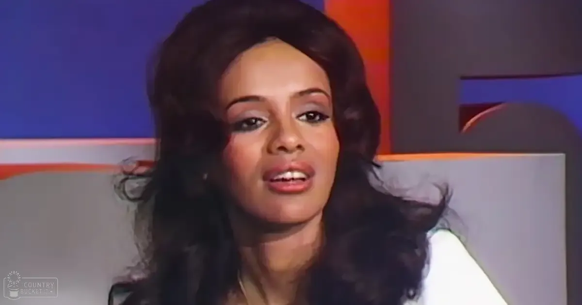 Young adult female with dark wavy hair indoors wearing a white high-necked garment against colorful geometric shapes in an evenly lit studio setting.