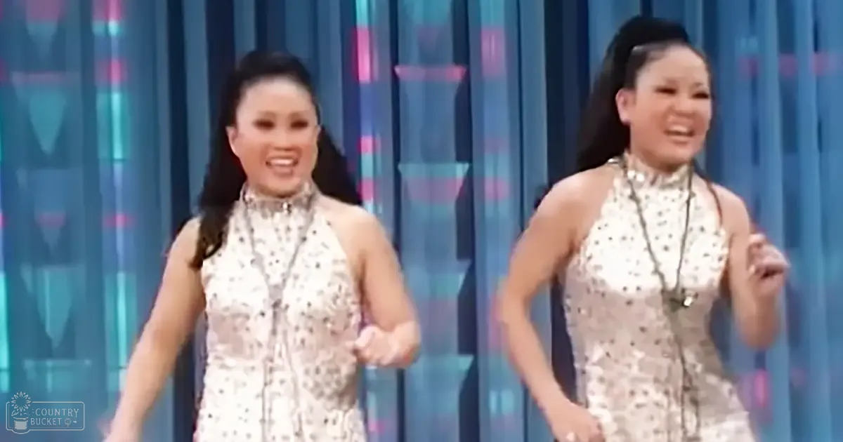 Two young adult female performers wearing sleeveless, shimmering silver sequined halter necklines on a stage with blue and pink vertical striped backdrop under bright lighting.