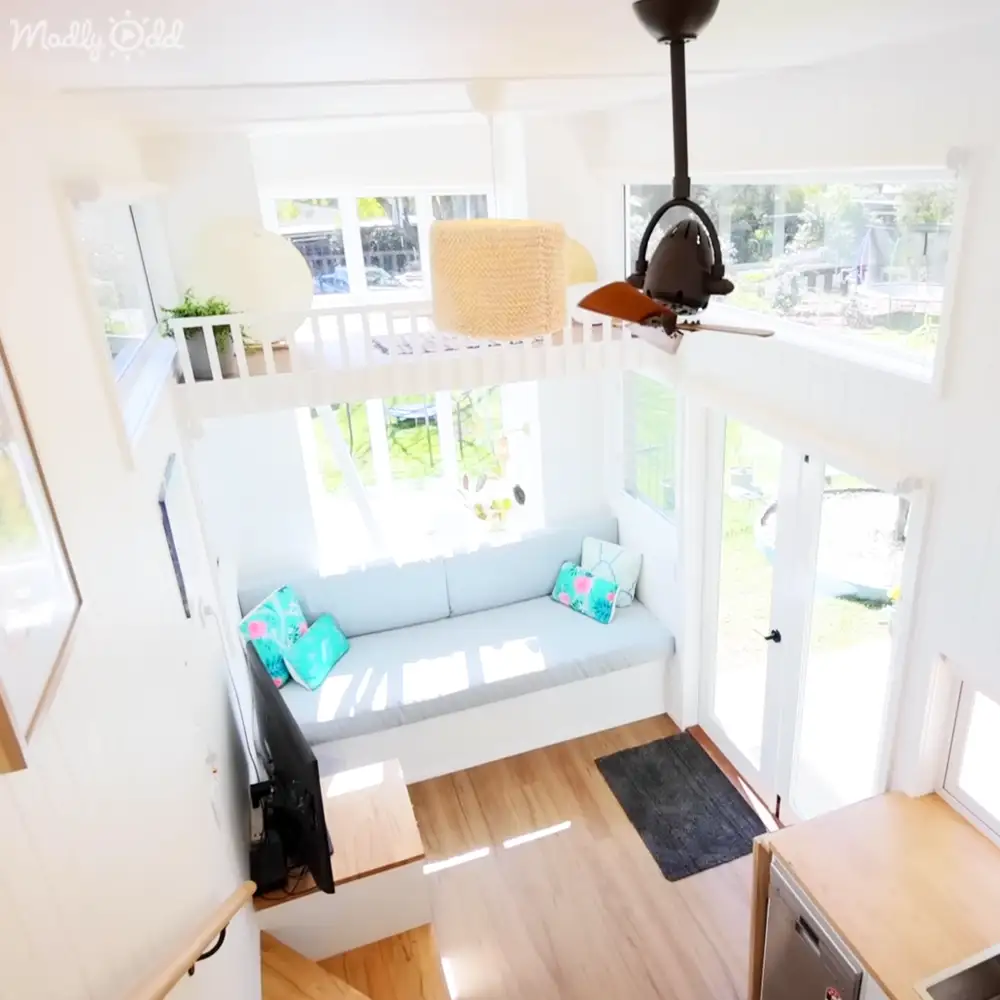A modern minimalist living space with light blue sofa, floral pillows, wooden floor, small rug, wall-mounted TV, ceiling fan, hanging lamp, and large windows showing clear daytime greenery.
