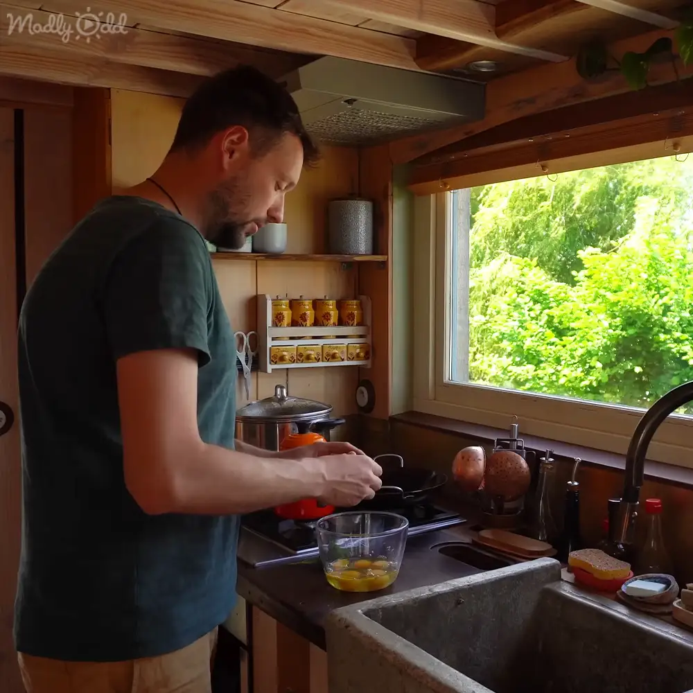 An adult male in a relaxed, casual dark green short-sleeved t-shirt stands in a cozy kitchen with wooden beams, jars of spices, and lush greenery outside on a bright day.