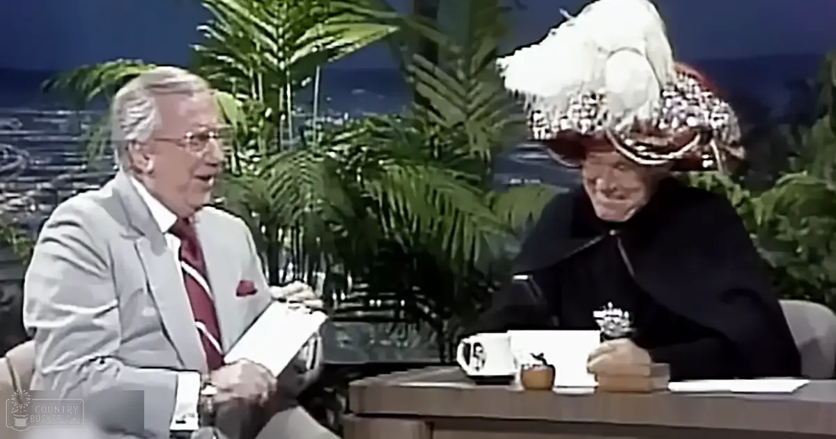 Two older adult males seated at a desk in an indoor studio with palm trees visible through the window and a large plant behind them.