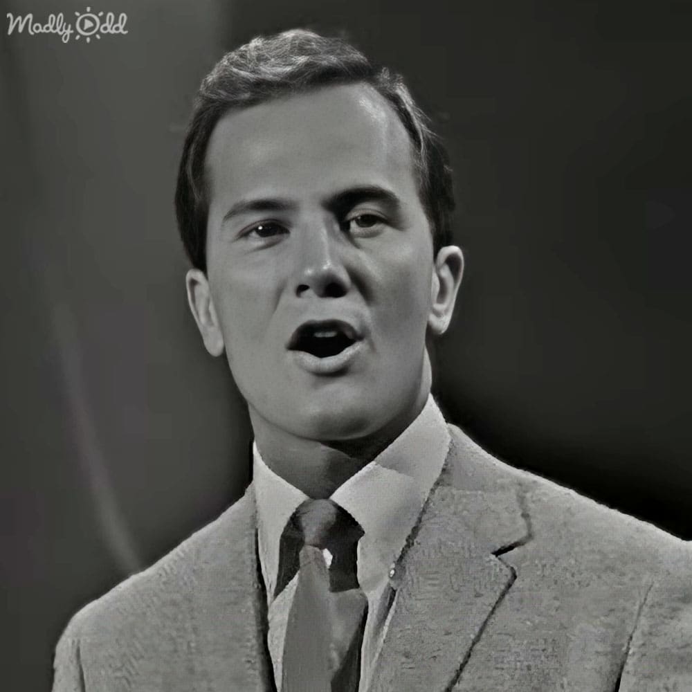 A young adult male wearing a tailored light gray suit jacket with a subtle pattern, standing against a dark gray backdrop in an indoor setting.