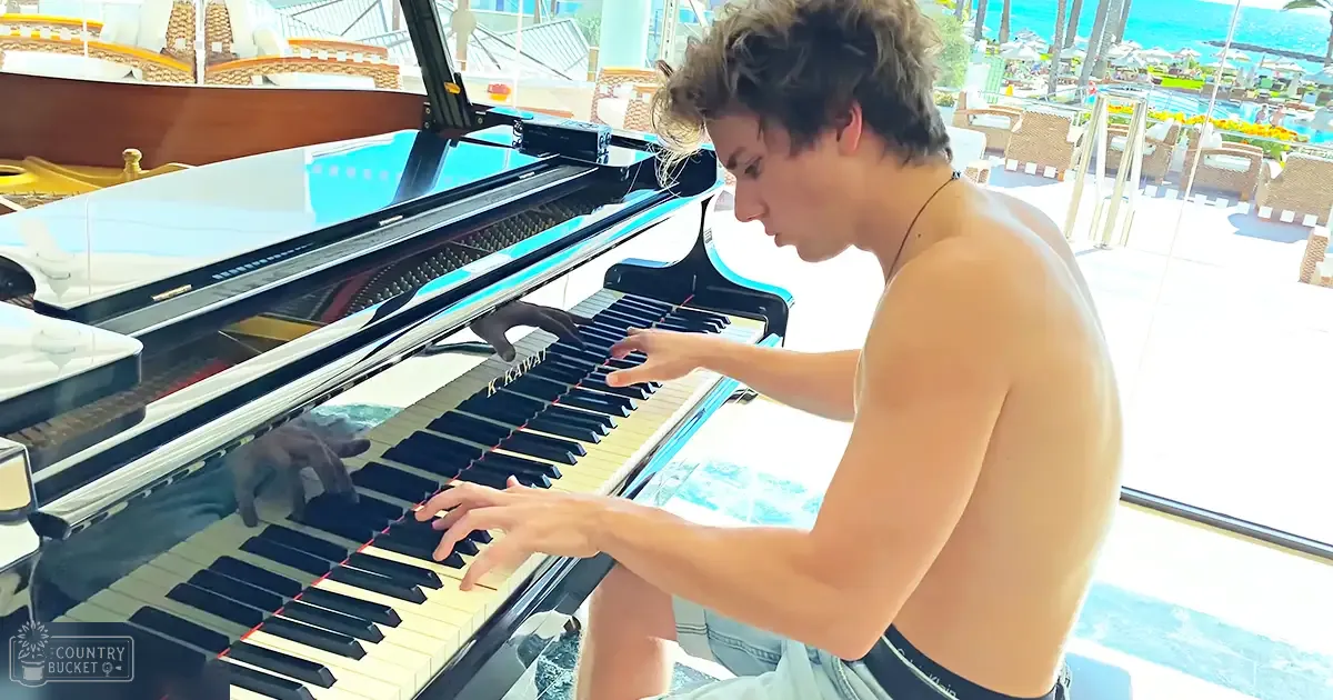 A young adult male shirtless, wearing light-colored jeans with a dark belt, plays a grand piano outdoors near a beach setting under clear skies.