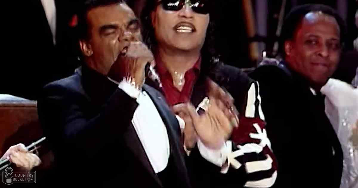 A middle-aged man in a black tuxedo jacket with white lapels is singing into a microphone at an indoor formal event, accompanied by other individuals in formal attire.
