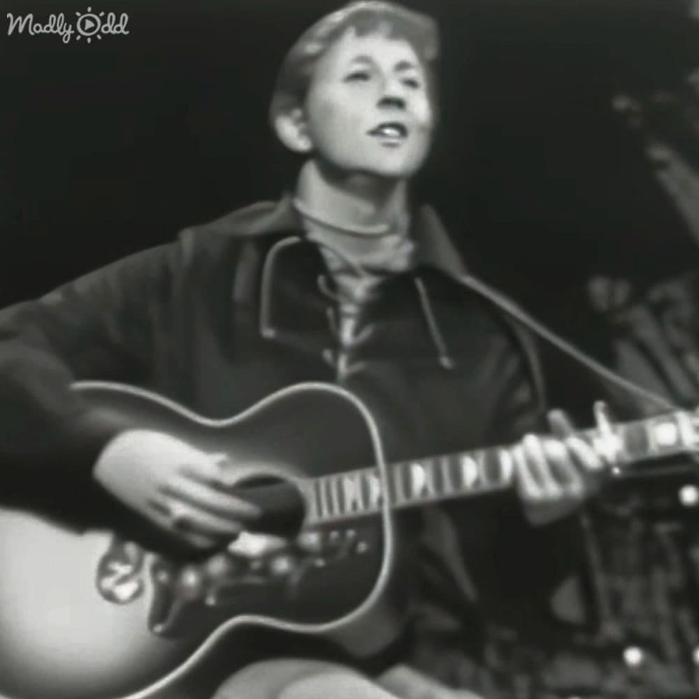 Young adult male playing guitar indoors with a microphone stand visible on the right.