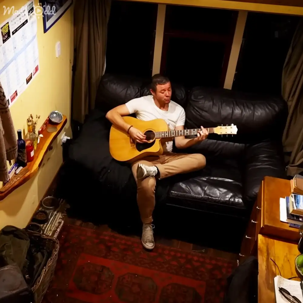 A young adult male plays guitar in a cozy living room with yellow walls, black leather couch, wooden shelves, and artificial lighting.
