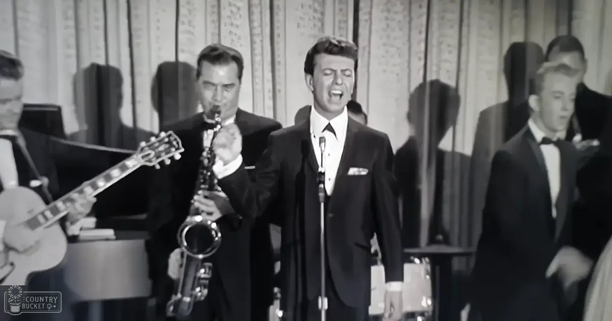 An adult male singer performing at a concert with an ensemble, surrounded by patterned stage curtains and microphones.