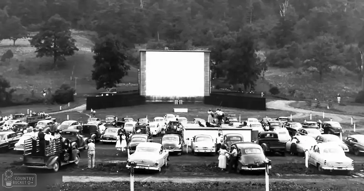 An adult male singer in a drive-in movie theater with older model cars parked in rows under natural daylight, set against an open field with trees in the background during spring or summer.