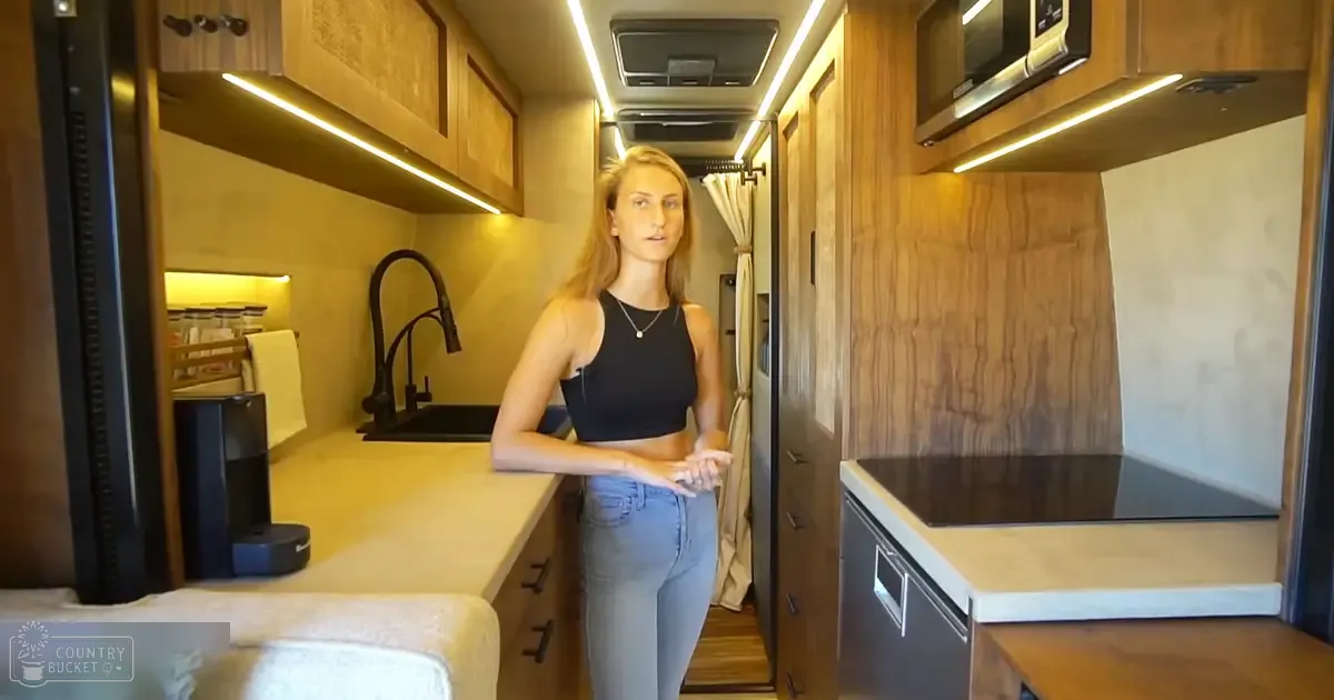 A young adult female in an RV interior with a black sleeveless crop top and light blue jeans.