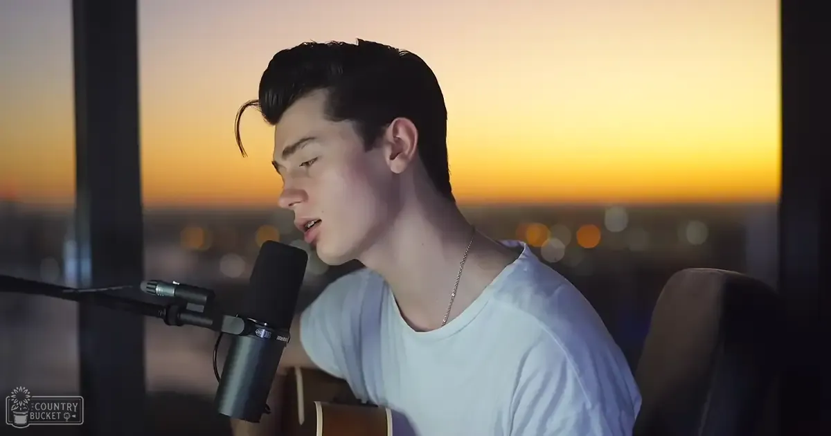 A young adult male wearing a light blue t-shirt sits indoors against a backdrop of an orange and yellow cityscape at sunset or sunrise, suggesting spring or fall.