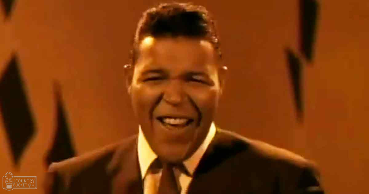 Middle-aged male wearing a dark suit jacket with 'Madly Didd' text in an indoor setting with warm orange tones and stage-like backdrop.