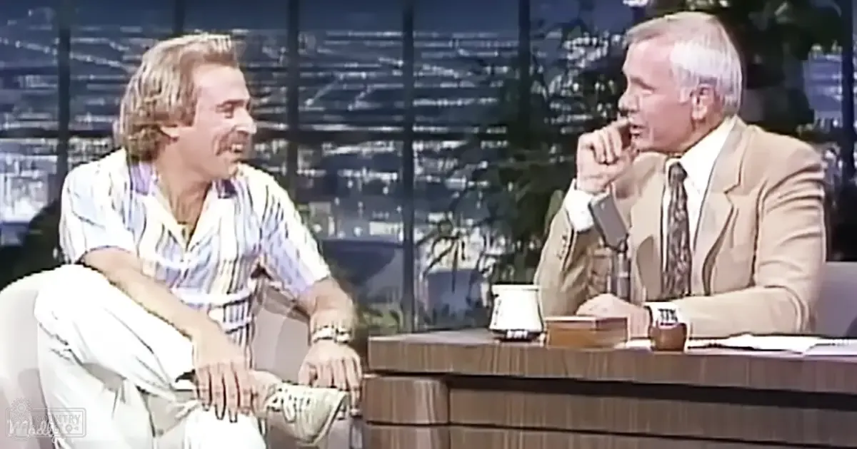 Adult male talk show host in a white and blue striped shirt interviews an older adult male guest in a beige suit jacket over a patterned tie, seated at a wooden desk with urban cityscape view through large windows in the evening.