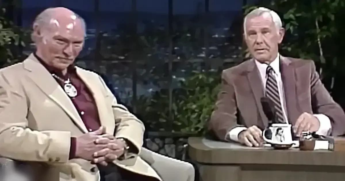 Two adults in a talk show setting with an older male wearing a beige blazer over a maroon patterned shirt, seated at a wooden desk, holding a mug and small items. A middle-aged to older adult male in a brown blazer and white striped tie sits beside him.