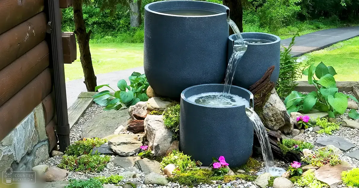 Three large black cylindrical pots arranged vertically as part of a water fountain system in an outdoor garden with lush greenery and pink flowers.