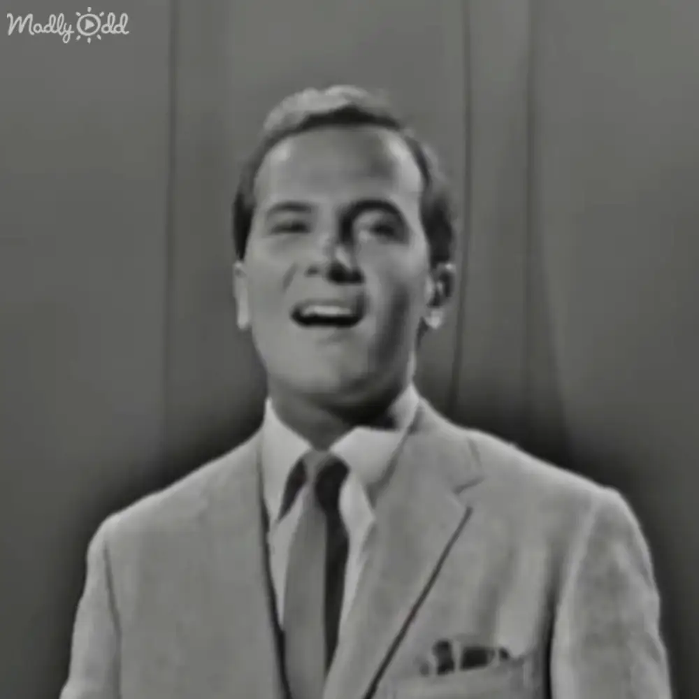 A middle-aged man in a light-colored suit jacket with a pocket square and dark tie stands against an indoor backdrop, possibly in a formal or professional setting.