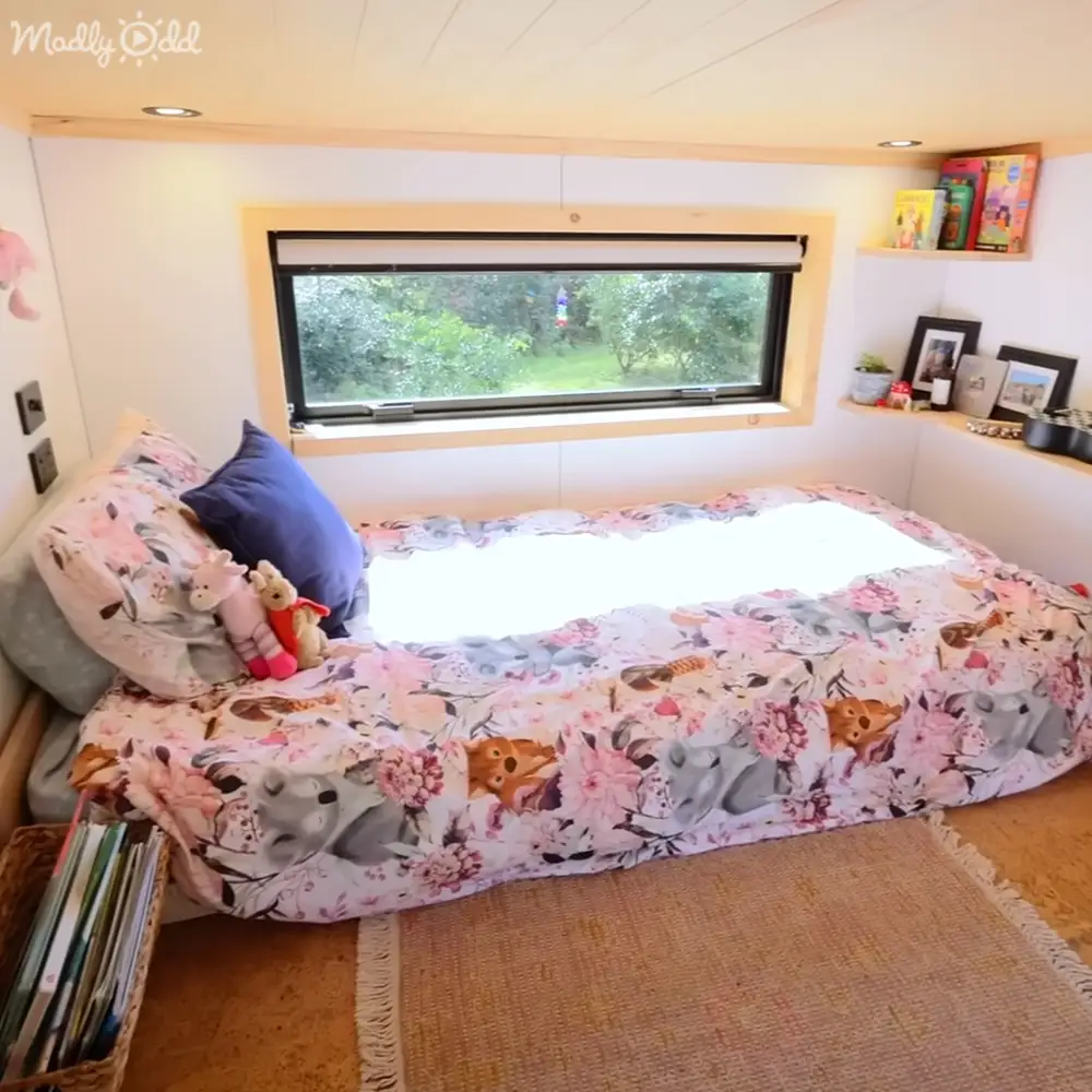 A cozy bedroom scene with a bed adorned with floral-patterned bedding with cartoon characters, a window showing greenery outside, and indoor plants and books on a shelf.