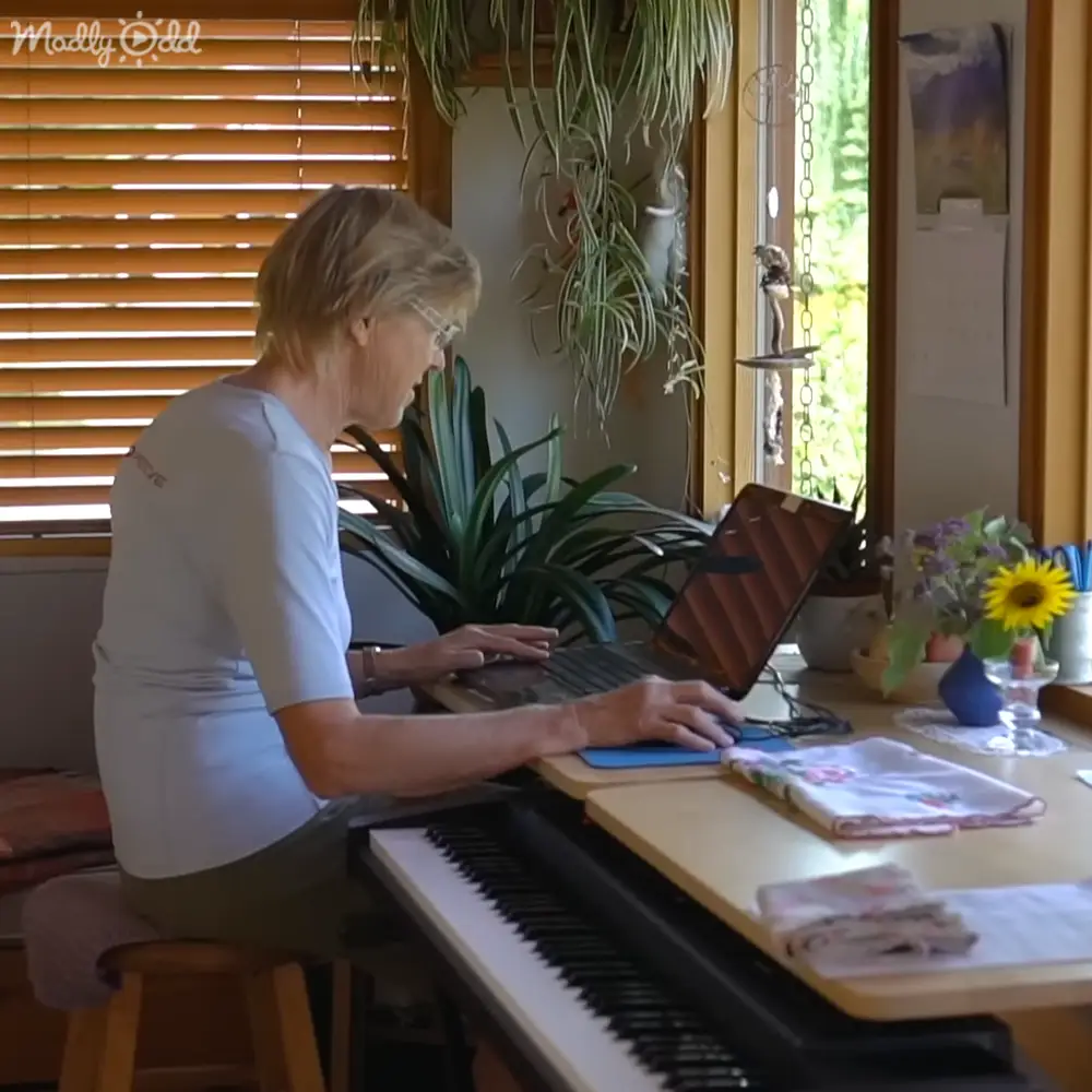 An older adult woman works at a wooden desk in her home office with natural light streaming through blinds and greenery around.