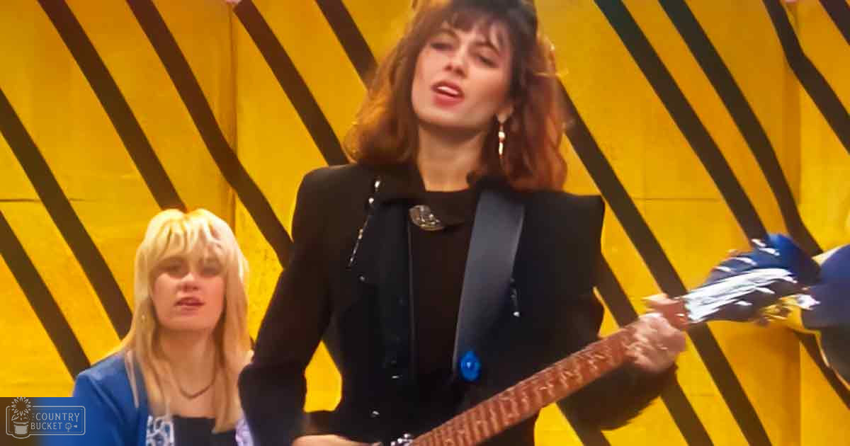 A young adult female playing an electric guitar, wearing a black jacket over a dark top and gold hoop earrings. Another individual in the background appears to be singing or speaking.