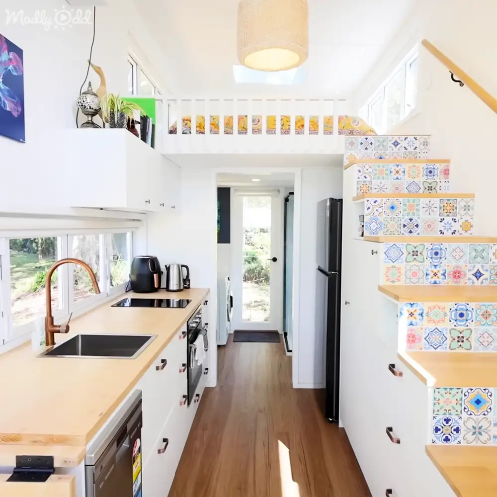 A modern kitchen with stairs leading up, located indoors; features a copper faucet over a sink, wooden countertops and cabinets, colorful decorative tiles on the staircase walls, black refrigerator, small oven or stove with kettle and other appliances, hanging light fixture above counter, artwork to left of sink.