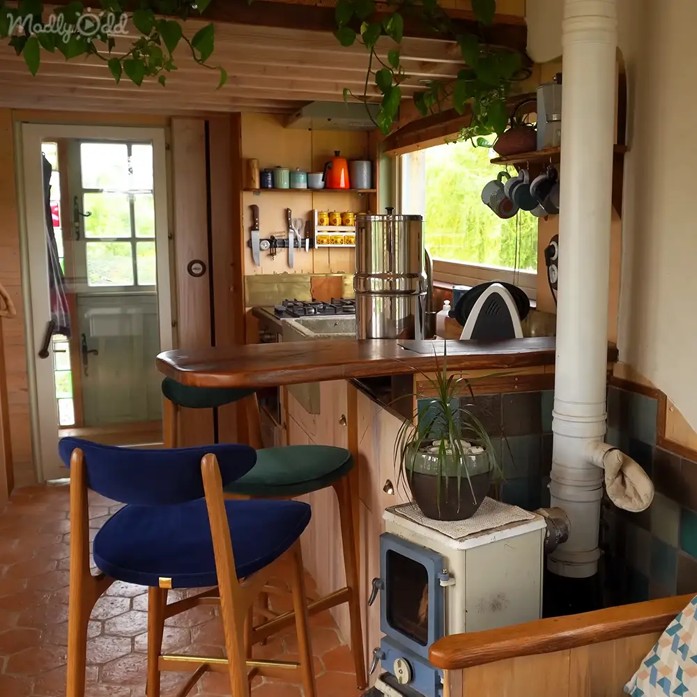 A cozy kitchen area with wooden bar stools, a small blue wood-burning stove, shelves holding pots and pans, and natural light streaming in from the window. The lush greenery outside suggests spring or summer.