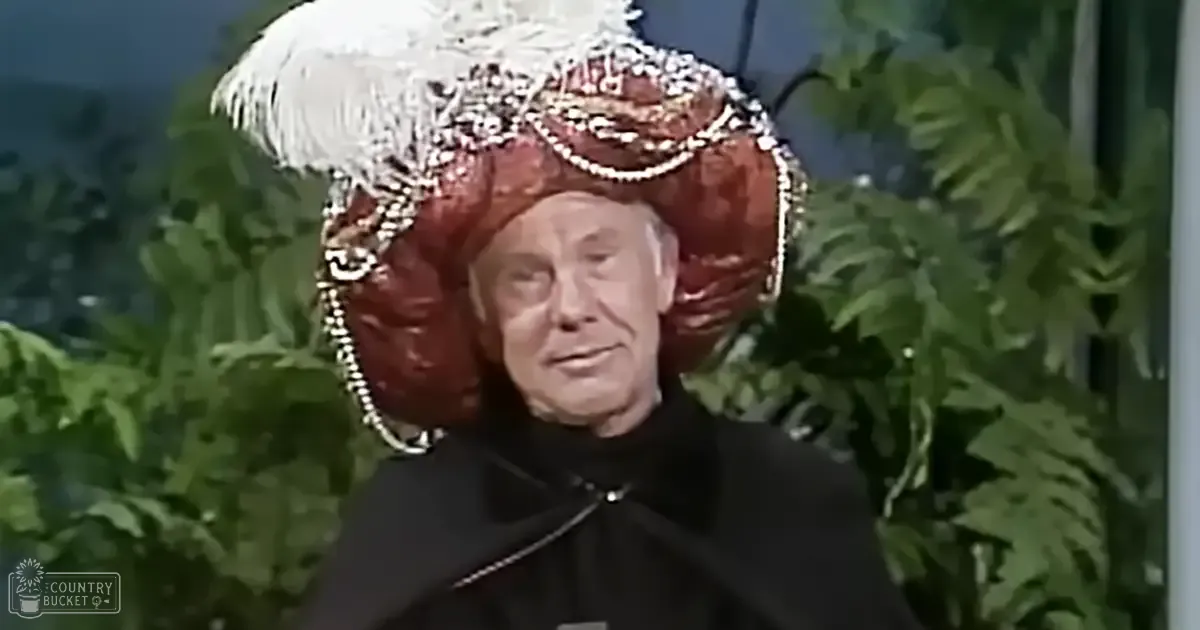 An older adult male wearing a black, high-collared robe with long sleeves stands in an indoor studio setting, surrounded by green foliage under controlled lighting conditions.