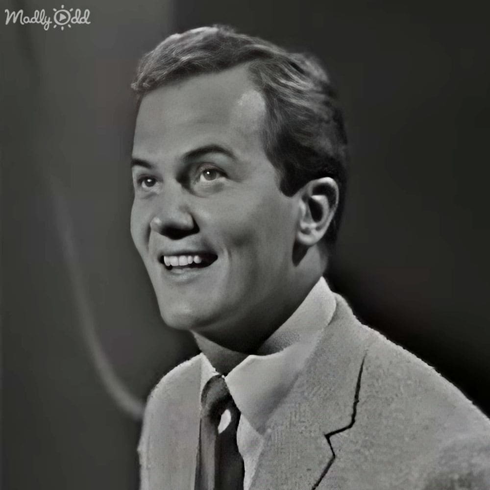 A young adult male wearing a light-colored suit jacket with a subtle pattern and a dark tie stands against a plain, dark backdrop in an indoor setting with soft lighting.
