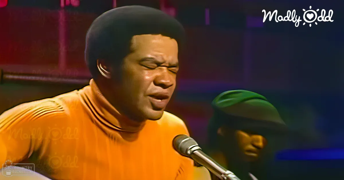 A male adult singer wearing a bright orange turtleneck sweater stands on what appears to be a stage in front of a microphone, with 'Madly Odd' text visible nearby.