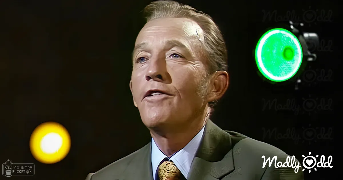 A middle-aged adult male wearing an olive green wool blend suit with a white collared shirt and patterned gold tie in an indoor studio or stage setting, illuminated by focused lighting.