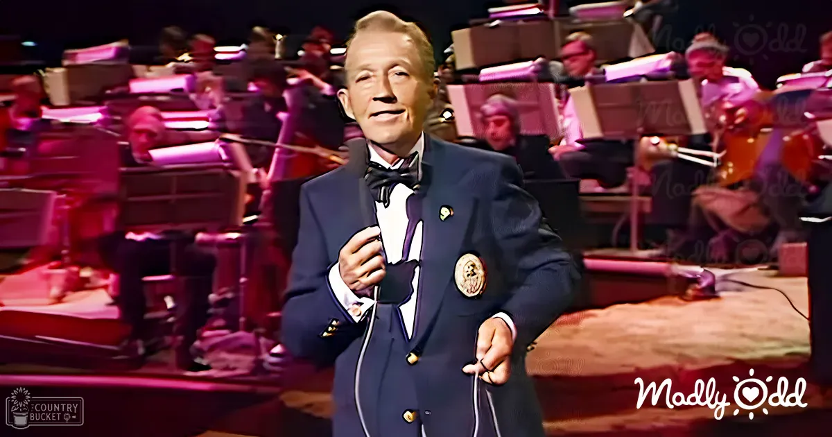 A middle-aged male performer wearing a dark blue double-breasted suit with white piping and gold buttons, standing before an orchestra in what appears to be an indoor concert hall at night.