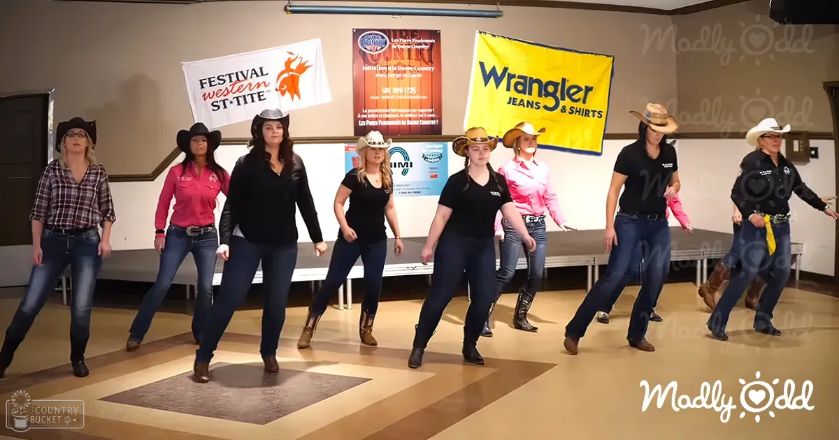 A group of young adult female dancers in casual attire perform at an indoor festival event, surrounded by promotional banners for Wrangler Jeans & Shirts and Festival Western St-Tite.
