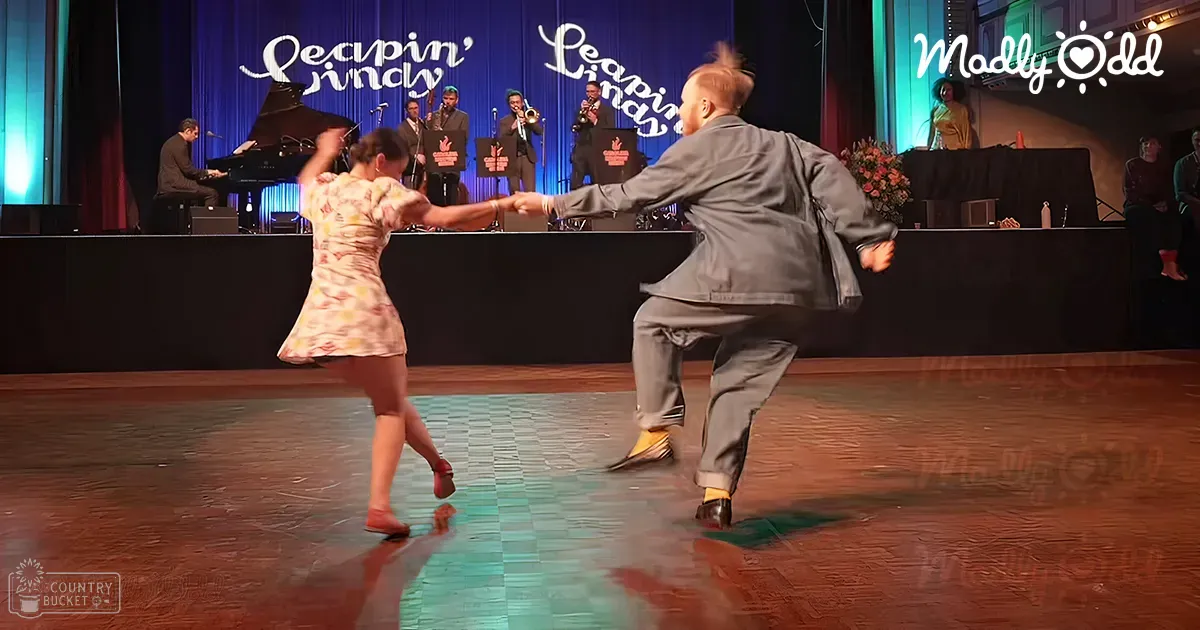 A young adult female dancer in a floral print knee-length dress with red shoes performs alongside a male dancer in a gray wool suit and yellow dress shoes on stage at 'Leavin' Lindy,' surrounded by band instruments, microphones, and a decorative backdrop.