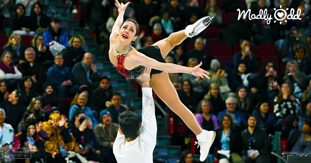 A young female figure skater in a black and red dress with sequins performs an aerial lift at a skating competition as spectators watch from rows of red stadium-style seats.