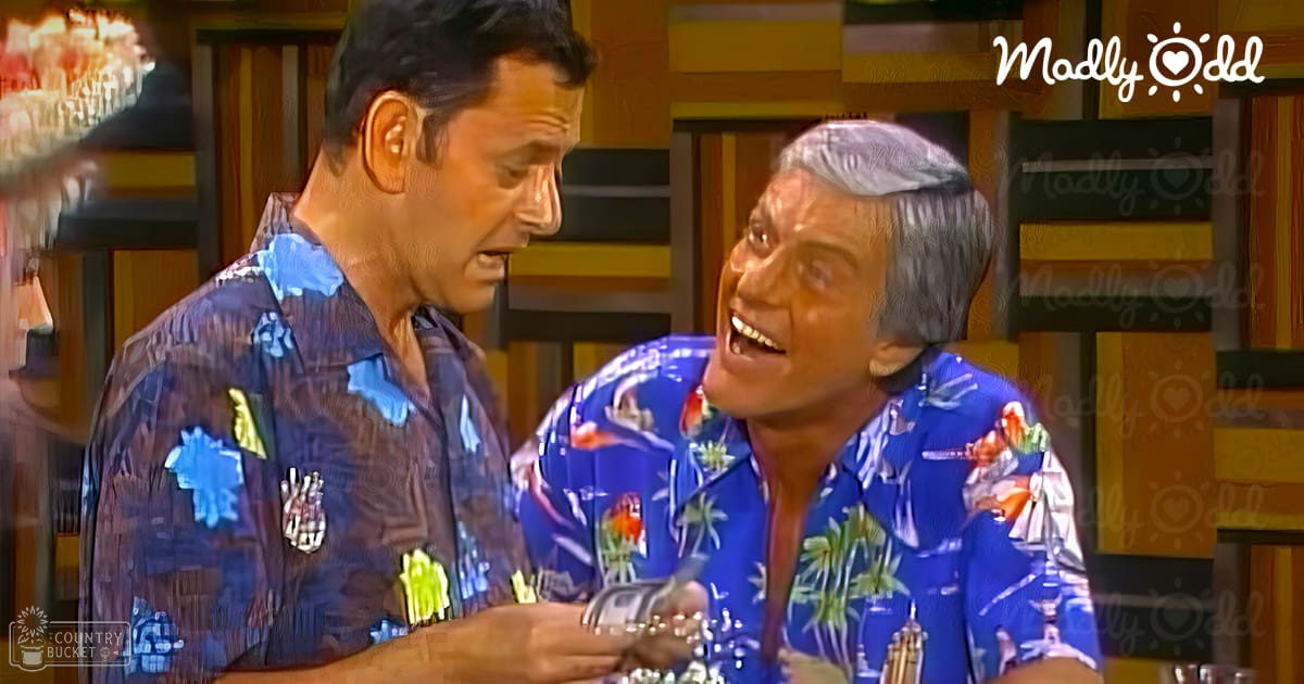 A comedic scene set in a casual indoor environment, with two adults wearing similar dark blue Hawaiian shirts with palm tree and tropical prints. The background includes a colorful geometric wall design and a partially visible bar with bottles and glasses.
