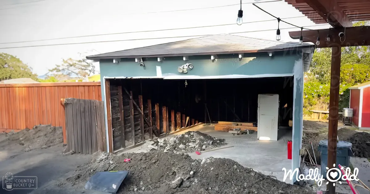 A residential backyard with a partially demolished wooden garage, dirt piles, outdoor string lights, red fire extinguisher, blue trash bin, and green trees under natural daylight, suggesting spring or summer.